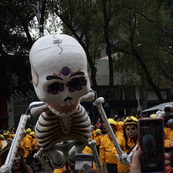 Una figura gigante de esqueleto se eleva sobre personas con cascos amarillos y rostros pintados, mientras varios asistentes toman fotos con teléfonos