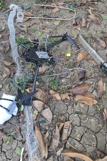 Dron de fibra óptica negro dañado en suelo seco y agrietado. Está enredado con una red blanca, rodeado de hojas secas y pequeñas plantas verdes