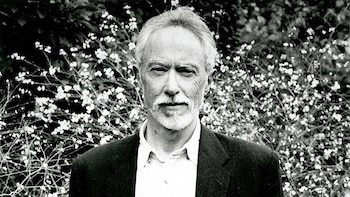 Retrato en blanco y negro del escritor John M. Coetzee con cabello y barba grises, mirando al frente, con un fondo de arbusto florido