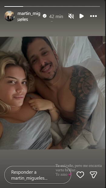 Martín Migueles y Wanda Nara, sonrientes, acostados juntos en una cama. Él está sin camiseta con tatuajes en el brazo, ella en una camiseta de tirantes gris