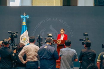 Una mujer en chaqueta roja habla en podio de madera. Bandera de Guatemala y fondo oscuro con texto "Gobierno de la República". Periodistas con cámaras