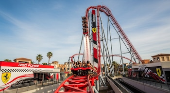 Montaña rusa Red Force con tren lleno de pasajeros ascendiendo una curva vertical roja con efecto de pulverización; edificios y palmeras bajo un cielo azul.
