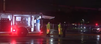 Un camión de soporte con luces rojas y compartimentos abiertos junto a rescatistas con abrigos fluorescentes en el pavimento mojado de noche