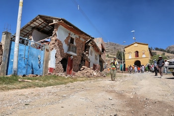 Una calle de tierra con dos casas al borde de la destrucción, sus paredes de ladrillo expuestas y escombros, mientras varias personas caminan bajo un cielo azul
