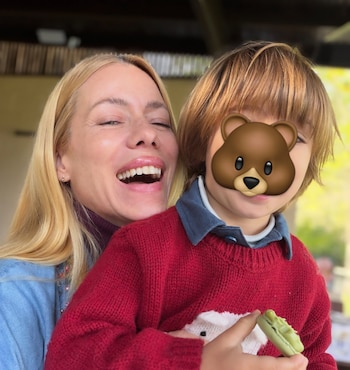 Una mujer rubia sonriente abraza a un niño con suéter rojo y el rostro cubierto por un dibujo de oso que sostiene un dulce verde