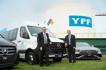 Dos hombres de traje posan en un área verde con tres vehículos Mercedes-Benz, incluyendo una Sprinter blanca, y un tanque de YPF al fondo