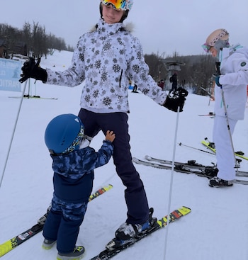 Tres esquiadores, dos adultos y un niño, equipados con cascos, chaquetas, esquís y bastones, sobre una pista de nieve con árboles al fondo