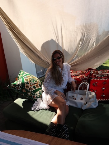 Una mujer con vestido de encaje blanco, gafas de sol y botas negras posa en un sillón verde junto a un bolso blanco