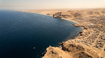 Vista aérea de la costa desértica peruana junto al océano Pacífico, con acantilados, pequeñas embarcaciones y un poblado adyacente