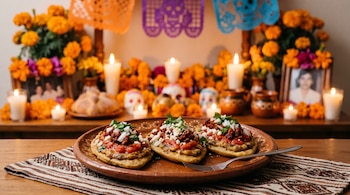 Tres tlacoyos con salsa, queso y chorizo sobre un plato de barro, acompañados al fondo por velas encendidas, flores anaranjadas y papel picado.