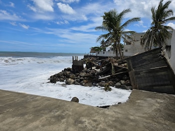 Fuerte oleaje rompiendo contra una costa rocosa y estructuras de concreto derrumbadas. Palmeras y edificios blancos se ven al fondo bajo un cielo azul