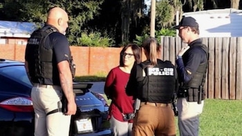 Tres agentes de policía con chalecos tácticos rodean a una mujer con gafas de ver junto a la parte trasera de un automóvil negro