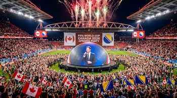 Estadio lleno de espectadores de noche con fuegos artificiales. En el centro, un escenario esférico muestra a Michael Bublé cantando, entre banderas de Canadá y Bosnia.