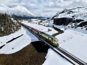 Tren verde, amarillo y rojo sobre un puente de ferrocarril. Paisaje montañoso con nieve, árboles y un río. Cielo con nubes blancas
