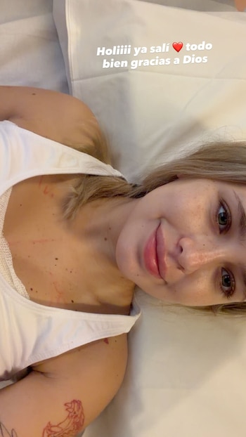 Mujer rubia en camiseta blanca acostada en una cama, tiene pecas y marcas rojizas en el pecho, un tatuaje en el brazo y texto superpuesto con un corazón