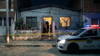 Una patrulla policial con sirenas rojas y azules encendidas está estacionada frente a una modesta vivienda acordonada con cinta amarilla de "POLICÍA - NO PASE".