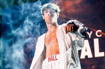 Bryce Hall sin camiseta y con una bata blanca exhala humo sobre un escenario iluminado con las letras de su nombre de fondo