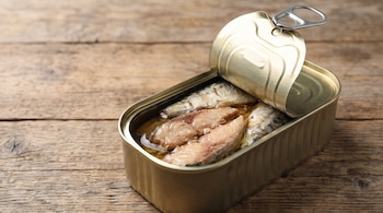 Lata de metal abierta con pescado enlatado y aceite. Se observa la tapa abierta con anilla y una superficie de madera debajo.