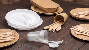 Platos, vasos y cubiertos de plástico blanco. Platos, vasos, envases y cubiertos de madera y papel marrón. Todo sobre mesa de madera.