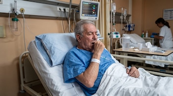 Un hombre mayor con bata azul y cánula de oxígeno tose en una cama de hospital frente a un monitor de signos vitales.