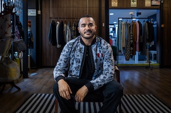 Un hombre con barba y una chaqueta de estampado de sellos de pasaporte y bandera peruana está sentado en una silla dentro de una tienda de ropa