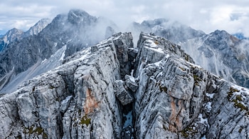 Vista aérea de una grieta profunda en la cumbre rocosa de una montaña con parches de nieve y niebla.