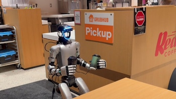 Robot humanoide bípedo plateado y negro con detalles azules luminosos en la cabeza, sosteniendo dos tazas de bebida, una verde y una negra, en un área de servicio