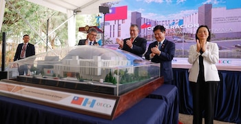 Cuatro personas con traje aplauden frente a una maqueta arquitectónica de un hospital protegida por una cubierta transparente