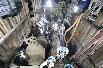 Trabajadores con trajes de protección blancos y cascos azules operan sobre tuberías industriales dentro de una excavación profunda