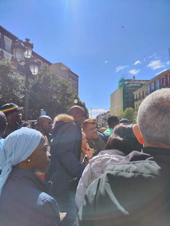 Grupo de personas, incluyendo Serigne Mbaye, apiñadas al aire libre bajo un cielo azul claro con sol brillante. Edificios y árboles verdes se ven al fondo