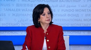 Mujer de cabello oscuro con chaqueta roja y botones dorados, sentada frente a una mesa con una tablet y un bolígrafo. Fondo azul con texto internacional