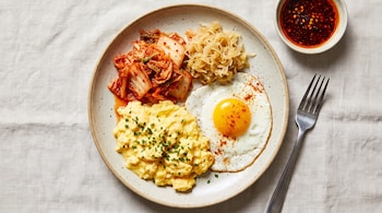 Plato con huevos revueltos, huevo frito, kimchi, chucrut, cuenco con salsa picante y tenedor sobre mantel de color claro.
