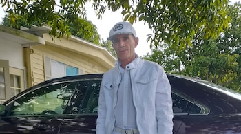 Hombre de gorra y chaqueta blancas de pie ante un automóvil oscuro y una casa amarilla de madera bajo la sombra de unos árboles.