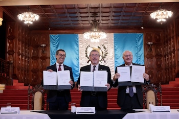 Tres hombres de traje sostienen documentos abiertos frente a una bandera de Guatemala; hay carteles con sus nombres, paredes de madera y lámparas colgantes
