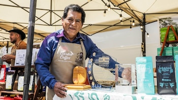 Un hombre con delantal sostiene trofeos y un reconocimiento junto a paquetes de café sobre una mesa en un puesto de feria