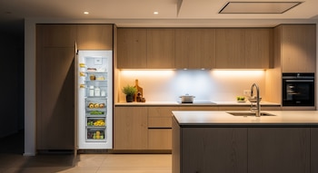 Cocina moderna con gabinetes de madera clara y nevera empotrada abierta. Se ven la encimera blanca, fregadero, horno y placa de inducción. Iluminación cálida.