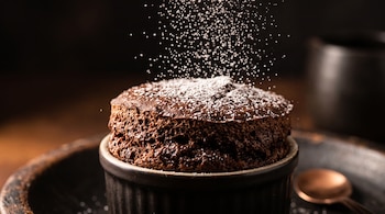 Soufflé de chocolate en un recipiente cerámico oscuro con partículas blancas de azúcar que caen sobre la superficie.