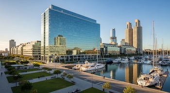 Vista aérea de un gran edificio de oficinas de cristal azul junto a una marina con muchos yates y veleros. Hay otros edificios, un parque y gente caminando bajo un cielo despejado.