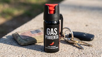 Un frasco de gas pimienta negro con tapa roja, con las palabras 'GAS PIMIENTA', un chile y un aviso, está sobre concreto junto a un monedero y llaves.