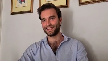 Hombre joven con barba, camisa de rayas azul claro, sonriendo a la cámara. Al fondo, una pared blanca con dos cuadros enmarcados dorados