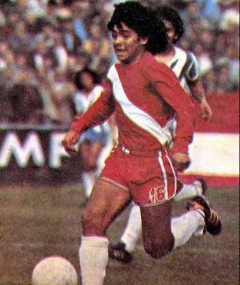 Un futbolista joven con uniforme rojo y franja blanca corre con un balón sobre una cancha de césped con público en la tribuna de fondo