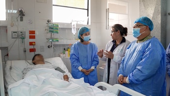 Cuatro personas en una habitación de hospital: un paciente en cama, una doctora y dos acompañantes con ropa quirúrgica y mascarillas faciales