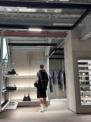 Un hombre de espaldas, con sudadera sin mangas negra y shorts, observa los productos exhibidos en estantes iluminados dentro de una tienda