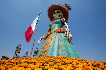 Figura de una catrina con vestido turquesa rodeada de flores de cempasúchil, junto a una bandera nacional y las torres de una catedral.