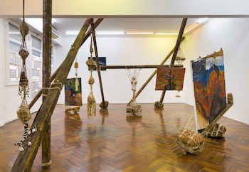 Estructura de madera, objetos trenzados suspendidos, pinturas, cuadros apoyados, piso de madera de parqué, paredes blancas, iluminación de galería