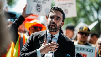 Zohran Mamdani, un hombre con barba oscura y traje, habla frente a micrófonos con su mano derecha levantada. Al fondo, personas y pancartas borrosas