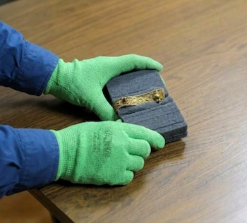 Dos manos con guantes verdes sostienen un bloque de espuma gris con una pieza dorada sobre una mesa de madera