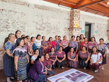 Grupo de aproximadamente 25 mujeres, muchas vistiendo trajes tradicionales coloridos, posan sonrientes frente a una pared de ladrillo blanco. Algunas de pie, otras agachadas, con un cartel en el suelo