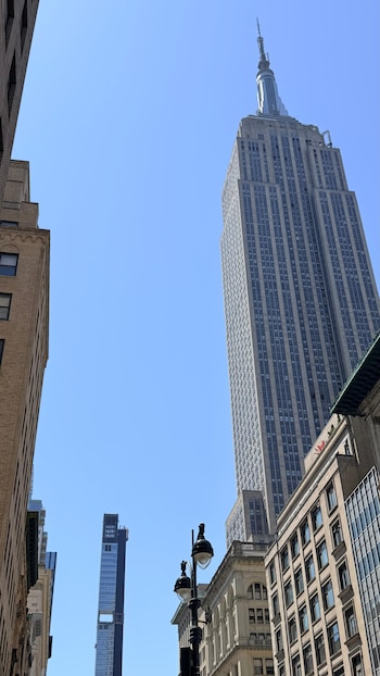 Vista ascendente del Empire State Building de Nueva York bajo un cielo azul claro, flanqueado por edificios; una delgada torre de cristal se ve al fondo