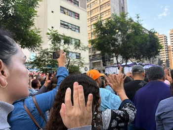 Personas de espaldas en una calle urbana con las manos levantadas y teléfonos móviles frente a edificios y árboles